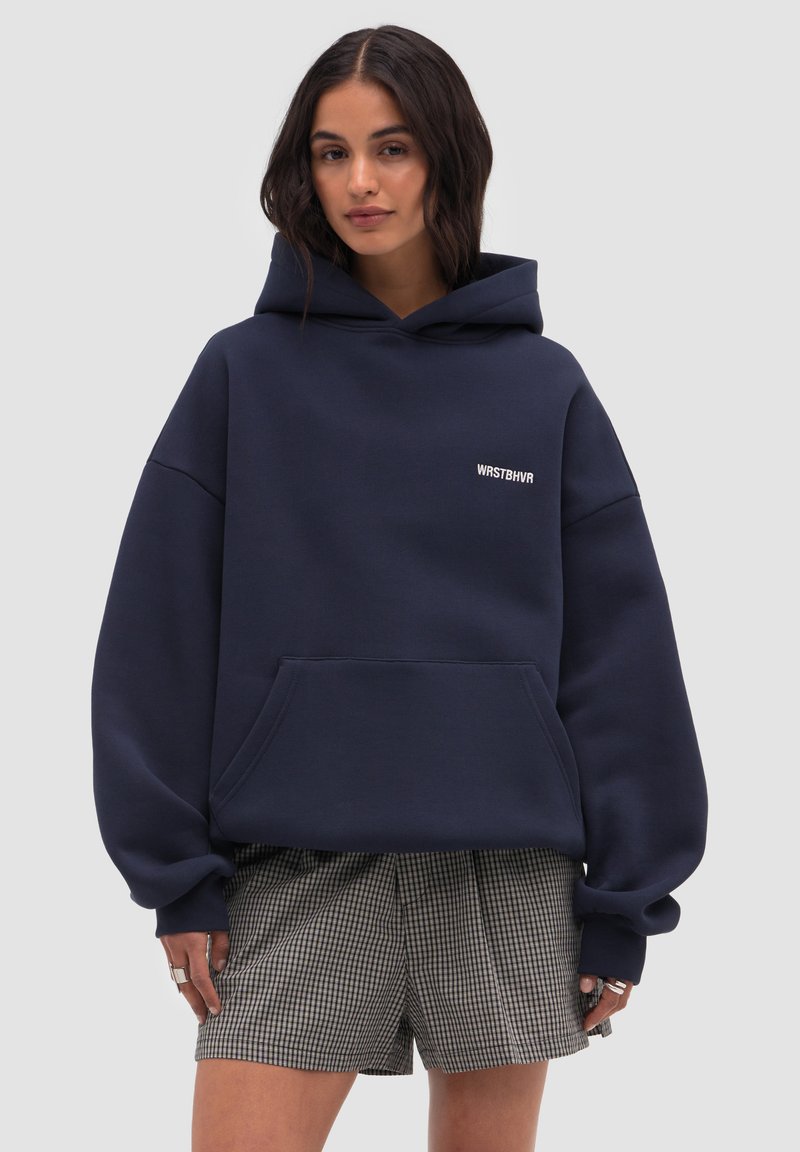 Vrouw draagt een oversized marineblauwe hoodie met de tekst "WRSTBHVR" en geruite korte broek, staand tegen een effen lichte achtergrond.