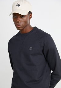 Felpa nera a girocollo con un logo discreto sul petto, abbinata a un cappellino da baseball color crema con un emblema più scuro.