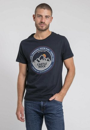 T-shirt à manches courtes bleu marine en coton, avec un design graphique rond représentant des montagnes et du texte en rouge et blanc. Associé à un jean bleu.