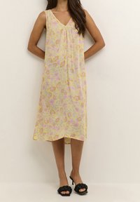 Kobieta w żółtej, kwiecistej sukience midi bez rękawów oraz czarnych, plecionych sanda�łach na obcasie, stojąca na tle jednolitego, jasnego tła.