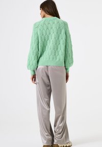 Minzgrüner Strickpullover mit einem strukturierten, gemusterten Design und überschnittenen Ärmeln, kombiniert mit hellgrauen Satin-Hosen und cremefarbenen Schuhen.