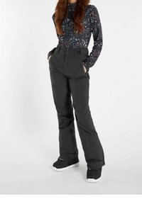 Kobieta w czarnych spodniach narciarskich i czarnych butach, z dłońmi w kieszeniach, w długim rękawie bluzce z galaktycznym wzorem, stoi na białym tle.