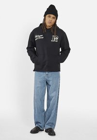 Czarna bluza z kapturem z białym tekstem i logo na piersi, zestawiona z jasnoniebieskimi szerokimi spodniami dżinsowymi i czarnymi butami. Modelka nosi czarną czapkę typu beanie.