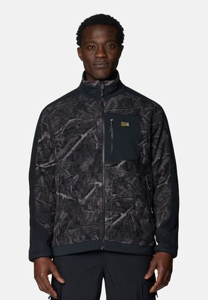 Mann trägt eine schwarz-grau gemusterte Fleecejacke mit Reißverschluss und Brusttasche und steht vor einem einfarbigen Hintergrund.