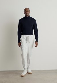 Μαύρο ζιβάγκο, άσπρα cargo παντελόνια με κορδόνι, και άσπρα sneakers με μπεζ σόλες, σε απλή ανοιχτόχρωμη φόντο.