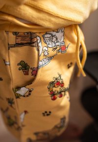 Gelbe Stoff-Jogginghosen mit Cartoon-Mustern, die Tiere, Pflanzen und Haushaltsgegenstände zeigen. Enthält einen Kordelzug und eine Seitentasche.