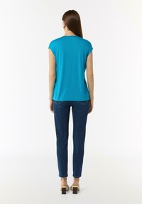 Türkises kurzärmliges Oberteil mit rundem Ausschnitt, kombiniert mit engen blauen Jeans. Der Stoff ist glatt mit einem leichten Glanz.