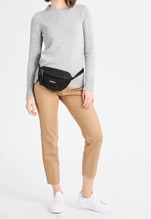 Persona vistiendo un suéter gris, pantalones cortos color canela, zapatillas blancas y una riñonera negra cruzada a la altura de las caderas sobre un fondo blanco liso.