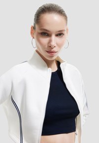 Weiße Zip-Jacke mit hohem Kragen und marineblauen Streifen an den Ärmeln, getragen über einem marineblauen Crop-Top. Verfügt über eine glatte Textur und taillierte Passform.