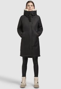 Schwarzer isolierter Mantel mit hohem Kragen und Fleecefutter. Verfügt über einen Frontreißverschluss, zwei Seitentaschen und einen geraden Saum. Getragen mit schwarzen Leggings und hellen Stiefeln.