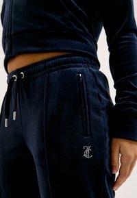 Ναυτικό βελούδινο φόρμα με φερμουάρ στις τσέπες, ελαστική μέση και λεπτομέρειες με ασημένιους θησαυρούς στο λογότυπο. Μαλακή υφή, εφαρμοστή σχεδίαση.