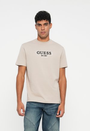 Joven con cabello corto y rizado, vistiendo una camiseta beige con el logo de GUESS y jeans azules, de pie frente a un fondo blanco liso.