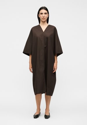 Γυναίκα στέκεται με το πρόσωπο προς τα εμπρός, φοράει ένα φαρδύ, σκουρόκαφέ φόρεμα μέχρι το γόνατο με φαρδιά μανίκια και μαύρα επίπεδα παπούτσια.