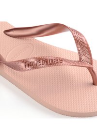 Tossut vaaleanpunaisesta kullasta, joissa on kuvioitu kumipohja ja yhteensopivat, joustavat hihnat, joissa on brändin "Havaianas" painatus sivussa.