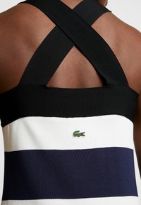 Vue arrière d'une personne portant un débardeur sans manches avec des bretelles croisées noires et des rayures horizontales bleu marine et blanches, présentant un petit logo de crocodile vert.