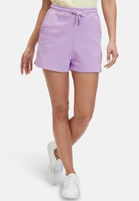 Femme portant un short à cordon lilas et des baskets blanches, debout jambes croisées sur fond blanc.