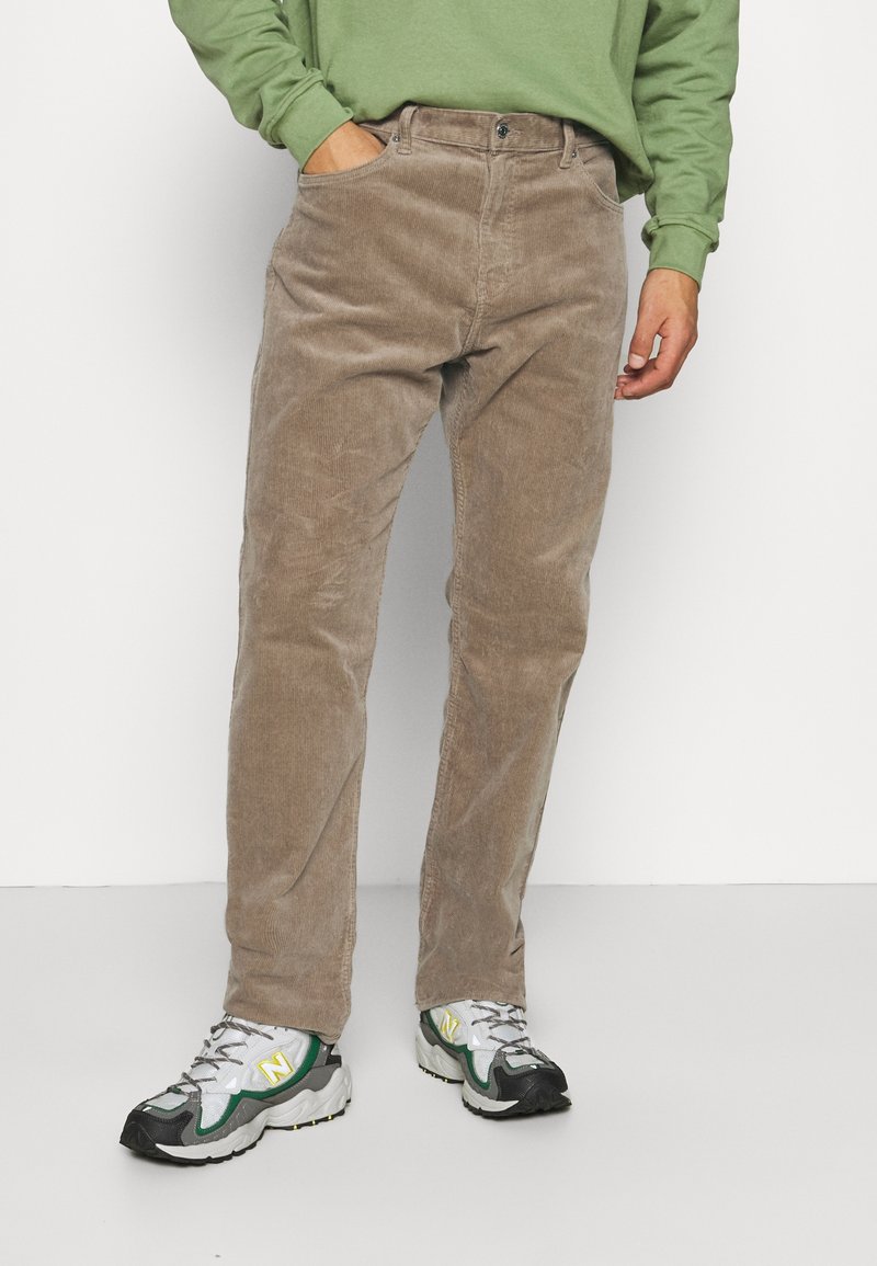 Pantalon en velours côtelé marron clair, doté d'une coupe droite, de poches avant et d'une texture subtile. Associé à des baskets grises et vertes.