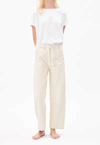 Personne portant un t-shirt blanc uni à manches courtes et un pantalon beige ample avec un lien à l'avant, debout pieds nus sur fond blanc.