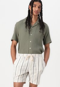 Chemise en jersey décontractée à manches courtes, de couleur vert olive, avec des boutons, associée à un short crème à rayures verticales bleues et une taille élastique.