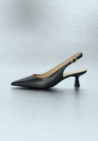 Chaussure slingback noire à bout pointu, talon bas et incliné, bride à boucle dorée, exposée sur une surface claire et réfléchissante.