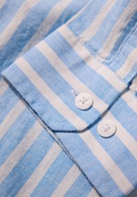 Collare di camicia a righe blu e bianche con due bottoni bianchi, realizzato in un tessuto testurizzato con una sensazione morbida e dettagli di cucitura visibili.