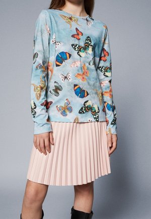 Kobieta ubrana w niebieską bluzkę z długim rękawem z kolorowymi nadrukami motyli oraz jasnoróżową plisowaną spódnicę, stojąca na tle szarego tła.