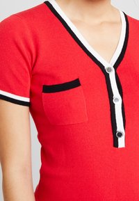 Haut en tricot rouge à manches courtes, col en V, bordure noire et blanche, avec une poche noire et des boutons brillants décorant le devant.