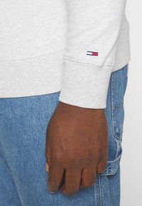 Felpa grigia con polsini a costine, con un piccolo logo ricamato in rosso, bianco e blu. Mano appoggiata contro jeans di denim blu.