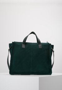 Tumma vihreä samettitote-bag, jossa on mustat nahkahihnat ja säädettävä olkahihna. Ominaisuuksiin kuuluu minimalistinen muotoilu ja jämäkkä muoto.