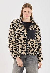 Fleece bunda v leopardím vzoru, béžová s čiernymi škvrnami, s čiernou zipsovou kapsou, vysokým golierom a voľným strihom. Nosená s šedými džínsami.