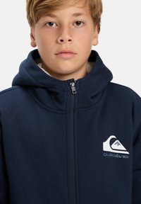 Sötétkék cipzáras kapucnis pulóver, húzózsinórral, puha belső résszel és hangsúlyos fehér Quiksilver logóval a mellkasán. Simább textúra.