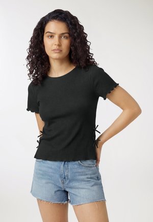 Donna con capelli ricci scuri che indossa una maglietta nera a maniche corte con fiocchi laterali e pantaloncini di jeans azzurri, in piedi con una mano sul fianco.