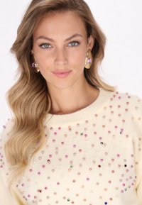 Pulover de culoare crem cu guler rotund, decorat cu mărgele multicolore. Păr ondulat care încadrează fața, având cercei mari din aur.