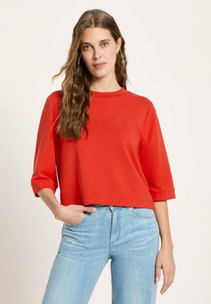 Young woman with long wavy hair wearing a red three-quarter sleeve top and light blue jeans, standing with one hand in pocket.
