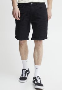 Homme portant un short noir retroussé, des chaussettes blanches Nike et des baskets noir et blanc, se tenant sur un fond blanc.