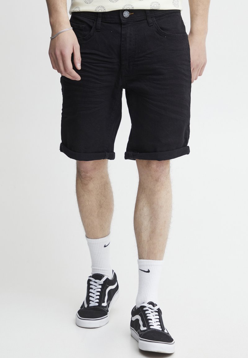Homme portant un short noir retroussé, des chaussettes blanches Nike et des baskets noir et blanc, se tenant sur un fond blanc.