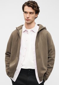 Καφέ φούτερ με φερμουάρ και υφή, φορεμένο πάνω από ένα ανοιχτό ροζ πουκάμισο με κουμπιά και λευκό μπλουζάκι. Το μοντέλο στέκεται μπροστά σε απλό φόντο.