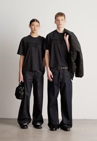 Doi modele poartă tricouri negre asortate, cu logo în relief, asortate cu blugi largi și de culoare închisă. Una ține o geantă mică neagră; cealaltă are o jachetă.