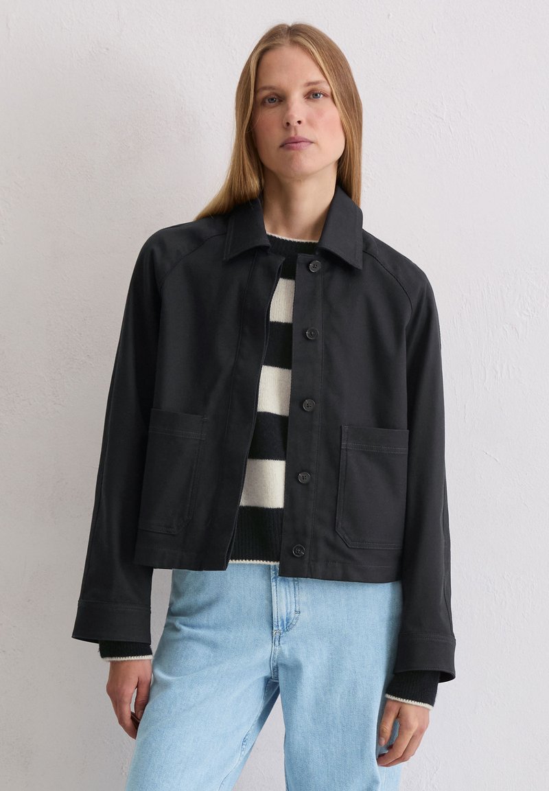 Czarna krótka kurtka z kołnierzem, zapięciem na guziki z przodu, dwoma kieszeniami i rękawami raglanowymi. W połączeniu z czarno-białym swetrem w paski.