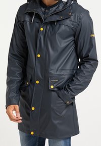 Parka imperméable marine avec capuche, dotée de boutons jaunes, de poches frontales et d'une finition lisse. Design classique avec intérieur rayé.