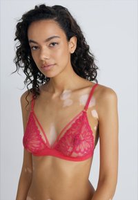 Soutien-gorge en dentelle rouge avec un motif floral, détails en maille transparente, bretelles ajustables et forme de tasse triangulaire. Le modèle présente le produit.