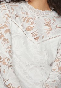 Top bianco in pizzo con intricati motivi floreali, caratterizzato da un colletto rotondo e un delicato bordo in pizzo sui lati. Tessuto morbido e semi-trasparente.