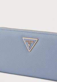 Gaiši zils teksturēts maks ar zelta trīsstūrveida Guess logotipu. Aprīkots ar rāvējslēdzēja aizdari gar augšējo malu. Gluda, izturīga materiāla.