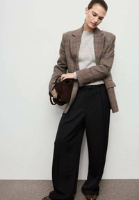 Blazer de cuadros marrón sobre un suéter gris, acompañado de pantalones anchos negros. Sostiene un pequeño bolso marrón oscuro. Textura suave, forma entallada.