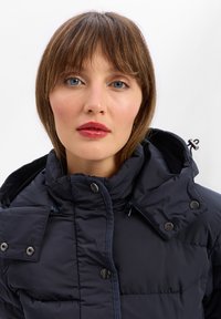 Granatowa kurtka puchowa z wysokim kołnierzem, zatrzaskami, zamkiem błyskawicznym i pikowanym wzorem. Posiada odpinany kaptur oraz regulowane sznurki.