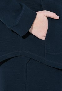 Top in tessuto blu navy testurizzato con polsino a coste e dettagli delle tasche laterali, caratterizzato da un orlo curvo e accenti di cuciture visibili.