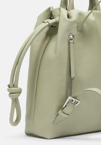 Sac en cuir vert clair avec une texture douce, doté d'une fermeture à cordon, d'une poche zippée à l'avant et de détails de sangle réglable.