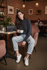 Kvinde i mørk sweater og lyse jeans, der sidder i en pink stol ved et cafébord og holder en hvid kop med en sort quiltet pung på bordet.