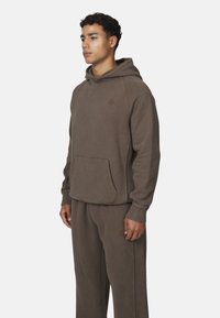 Felpa cappuccio marrone con tasca frontale, maniche raglan e logo testurizzato; abbinata a pantaloni felpati coordinati con strisce laterali.