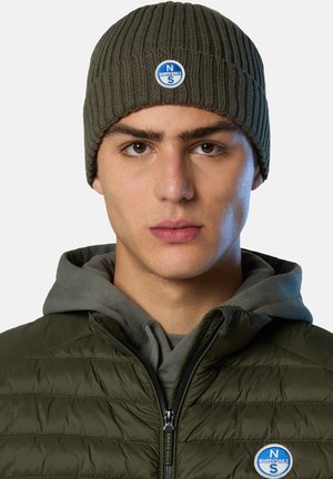 Olive green ribbed knit beanie with a blue round logo patch featuring "NORTH SAILS." Worn over a gray hoodie and quilted jacket.